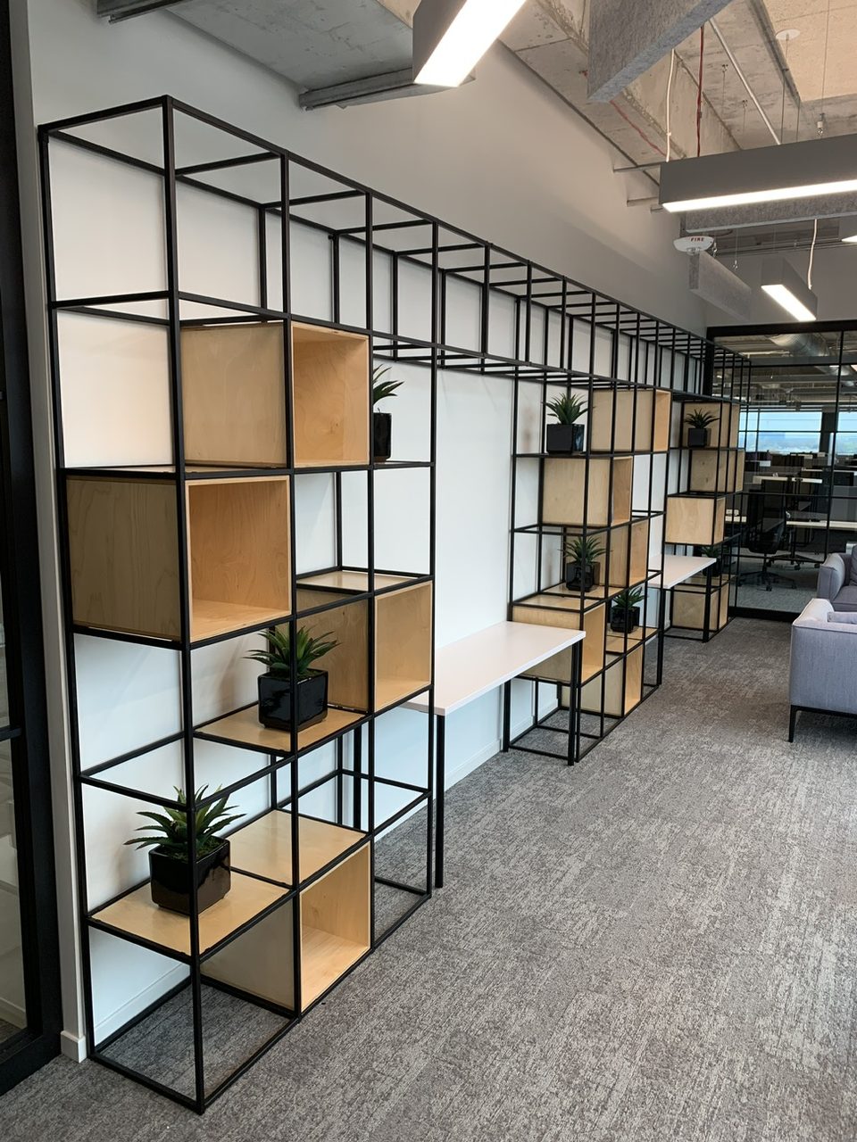 A black metal-grid panel-wall room divider with wood cubbies and plants in a finished office